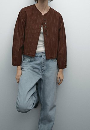 Personne portant une veste marron à rayures boutonnée sur un haut blanc, associée à un jean bleu clair taille haute, debout devant un fond uni.