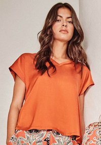 Kurzes, orangefarbenes Oberteil mit V-Ausschnitt, aus einem glatten Stoff gefertigt, mit lockerem Sitz und sanftem Fall am Saum.
