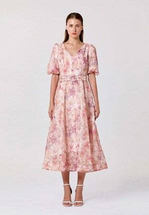 Femme portant une robe midi fleurie rose et beige avec des manches bouffantes, une taille cintrée avec ceinture et des talons blancs à bride à la cheville, debout devant un fond uni.