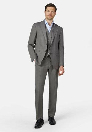 Man standing wearing a gray three-piece suit with a light blue shirt and black dress shoes on a white background.