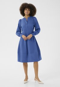 Blauwe jurk met lange mouwen, plooienrok, knoopsluiting bij de nek en een gestructureerde stof. Gekleed met beige hoge hakken.