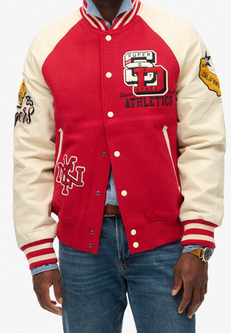 Homme portant une veste varsity rouge et crème avec des écussons sportifs, chemise bleu clair en dessous, jean bleu et montre-bracelet.
