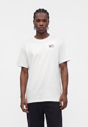 Jeune homme aux cheveux tressés, portant un t-shirt blanc Nike et un pantalon noir, debout devant un fond clair uni.