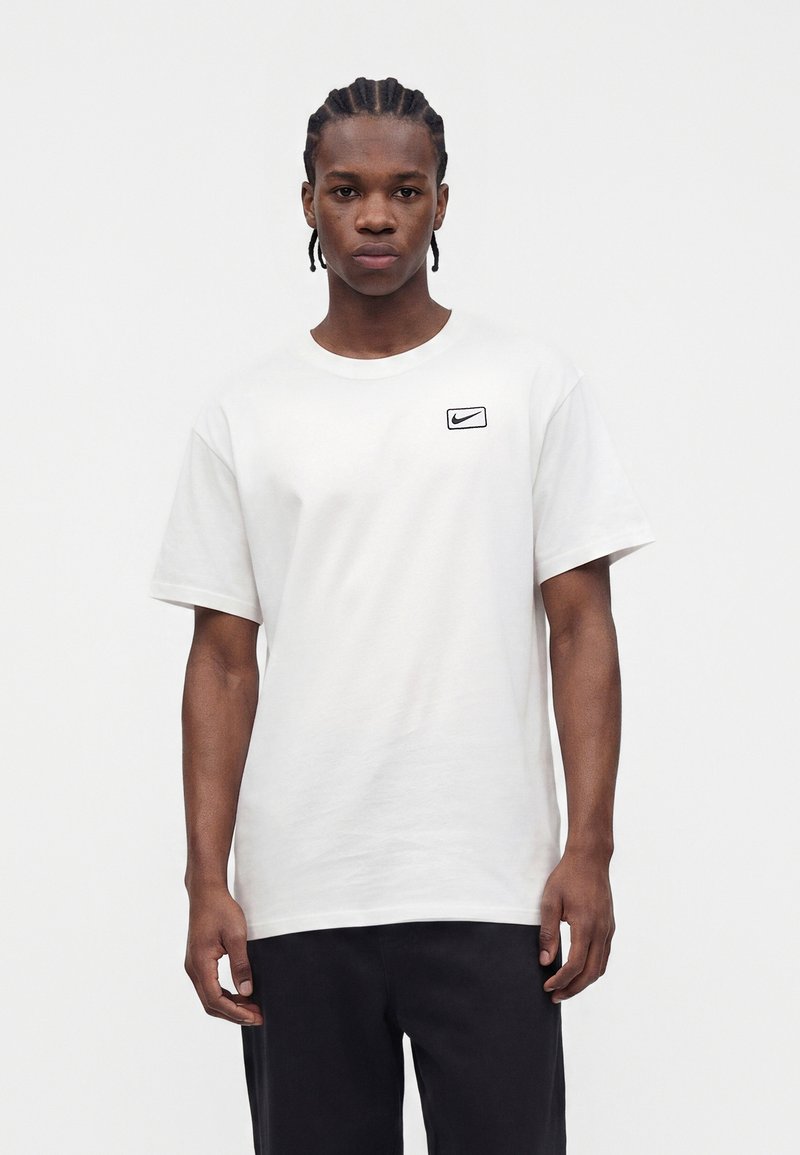 Junger Mann mit geflochtenem Haar, der ein weißes Nike-T-Shirt und schwarze Hose trägt, steht vor einem schlichten hellen Hintergrund.