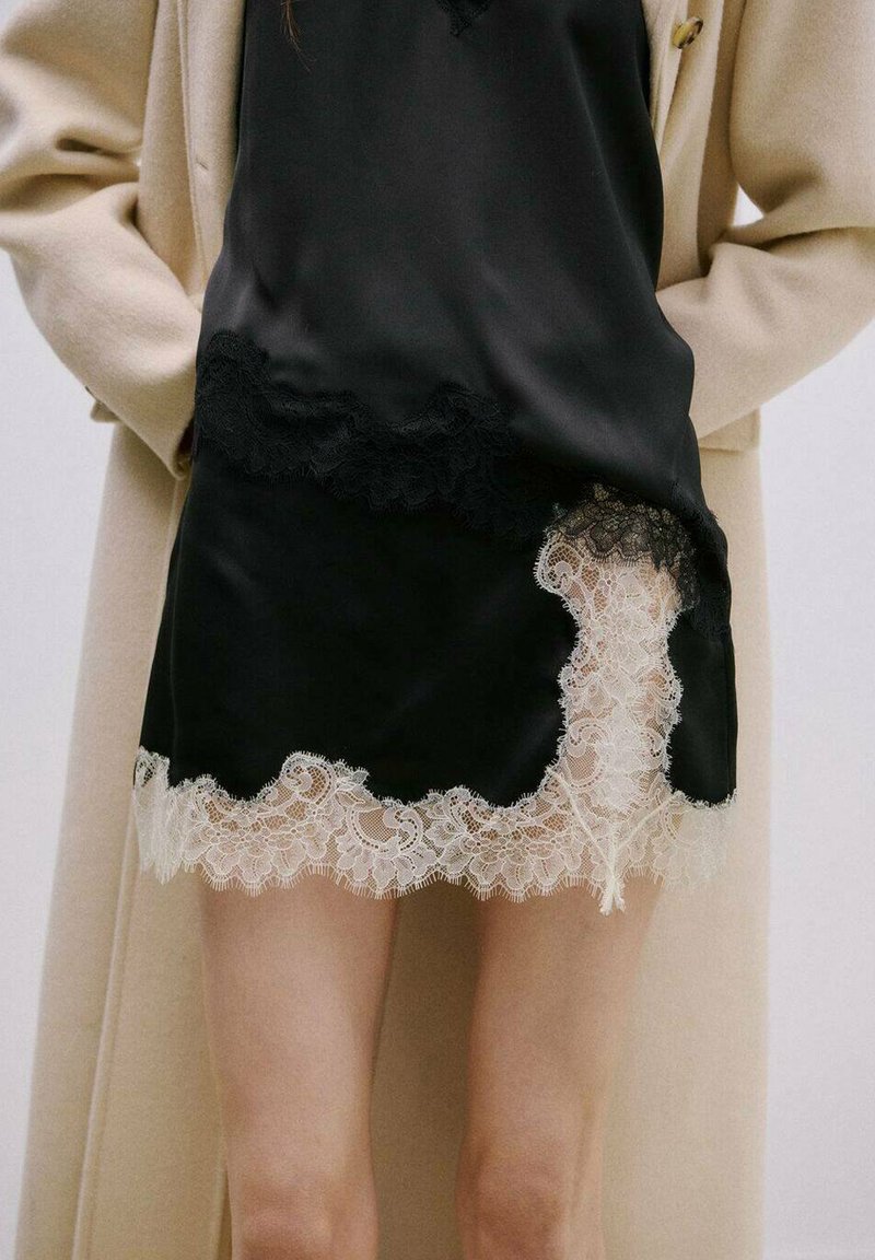 Femme portant une robe nuisette en satin noir avec un bord en dentelle blanche, un long manteau beige, les mains dans les poches du manteau, debout devant un fond uni.