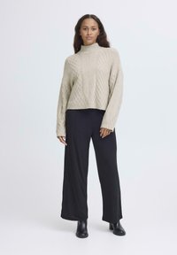 Maglione corto beige a maglia con colletto alto e motivo a diamante, abbinato a pantaloni neri a gamba larga e stivaletti neri.