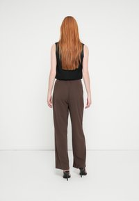 Črni zgornji del brez rokavov v kombinaciji s širokimi rjavimi hlačami. Hlače imajo gladko teksturo in rahle stranske zareze, ki jih dopolnjujejo črni čevlji s peto.