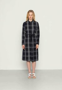 Vestido de manga longa, xadrez em azul-marinho com linhas brancas, cinto na cintura. Feito de um tecido com textura suave, até ao joelho. Usado com sandálias brancas.