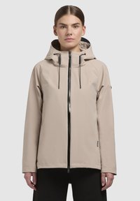 Beige vattentät jacka med hög krage, justerbar huva och svart dragkedja. Har raglanärmar och en minimalistisk design.