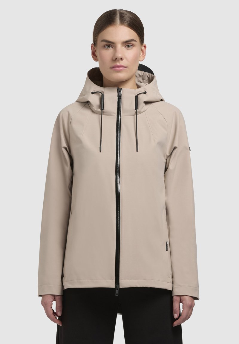 Beige vattentät jacka med hög krage, justerbar huva och svart dragkedja. Har raglanärmar och en minimalistisk design.