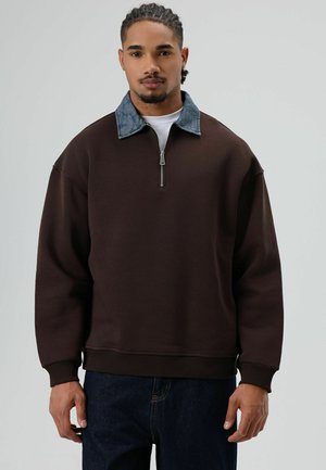 Bruine pullovertrui met een denim kraag, voorzien van een halve ritssluiting, geribbelde manchetten en een ontspannen silhouet, gecombineerd met donkere jeans.