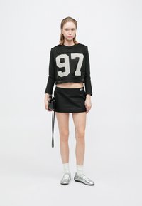 Schwarzes, kurzes Netzoberteil mit der weißen Zahl 97, kombiniert mit einem schwarzen Minirock. Das Model trägt silberne Schuhe mit weißen Knöchelsocken und hält eine kleine schwarze Tasche.