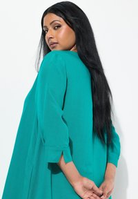Abito tunica in teal con maniche a tre quarti, caratterizzato da una vestibilità rilassata e spacchi laterali, realizzato in un tessuto leggero con una texture liscia.