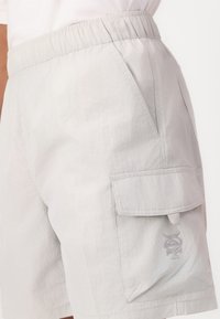 Bermudas gris claro con cintura elástica, bolsillos laterales y un bolsillo tipo cargo que presenta un sutil detalle de logo. Tejido ligero y texturizado.