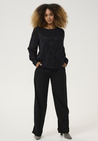 Blusa nera a maniche lunghe con motivo floreale e dettagli trasparenti, abbinata a pantaloni neri larghi. La modella indossa scarpe con tacco metalliche.