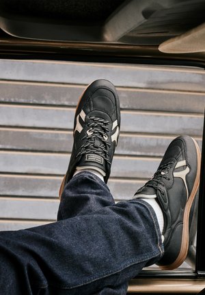 Persona che indossa scarpe da ginnastica bianche e nere e jeans blu con i piedi poggiati sul telaio della portiera di un’auto con pavimento grigio a righe all’esterno.