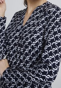 Navy blouse with a repeating white abstract chain pattern, featuring a V-neckline and long sleeves with buttoned cuffs. Smooth fabric texture.