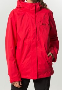 Veste imperméable rouge avec capuche, dotée d'une fermeture éclair frontale, d'une poche poitrine et de poignets élastiques. Fabriquée en matériau synthétique léger.