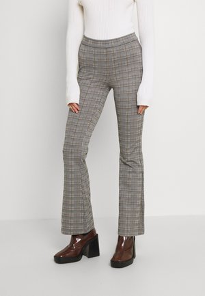 Vrouw met hooggetailleerde grijze geruite broek, witte geribbelde lange mouwen top en bruine enkellaarsjes met hak, staand op een effen achtergrond.