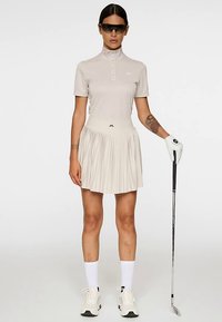 Vaaleanbeige golfpaita korkealla kauluksella, yhdistetty laskostettuun hameeseen. Asusteina valkoiset sukat, hansikas ja aurinkolasit. Kädessä golfmaila.