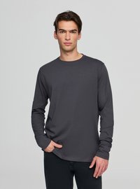 Ciemnoszara koszulka z długim rękawem wykonana z lekkiego materiału, z okrągłym dekoltem i dopasowanym krojem, noszona z czarnymi dżinsami.