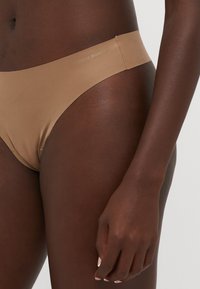 Beige naadloze bikini-onderbroek met een zachte textuur, voorzien van een brede elastische tailleband. De merknaam is subtiel op de tailleband gedrukt.
