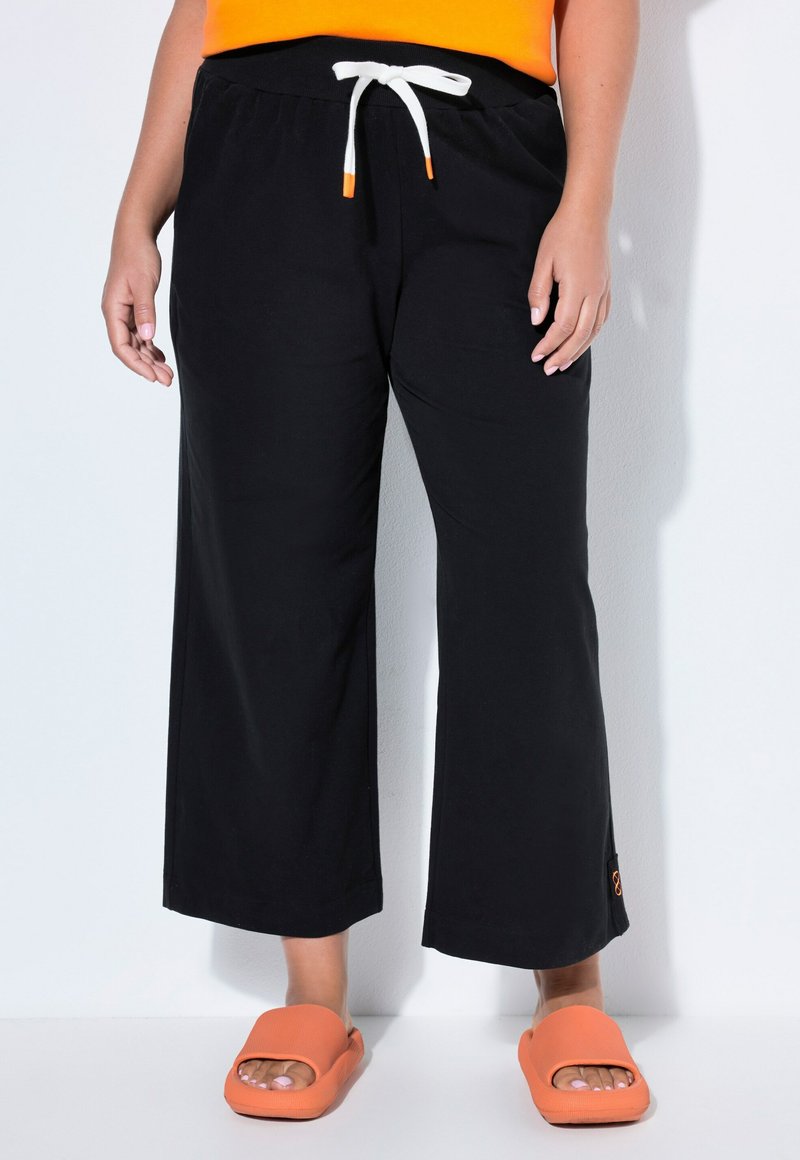 Persona che indossa pantaloni neri a gamba larga con cordoncino bianco e sandali arancioni slip-on, in piedi contro un muro bianco.