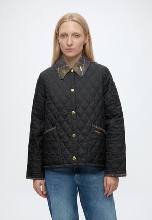 Femme aux cheveux blonds et raides porte une veste matelassée noire avec des boutons dorés, un col à carreaux et un jean bleu, debout devant un fond uni.