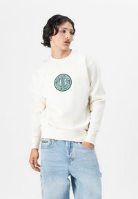 Felpa crema con un logo verde rotondo contenente del testo, abbinata a jeans baggy blu chiaro. Tessuto liscio, maniche lunghe e polsini a coste.