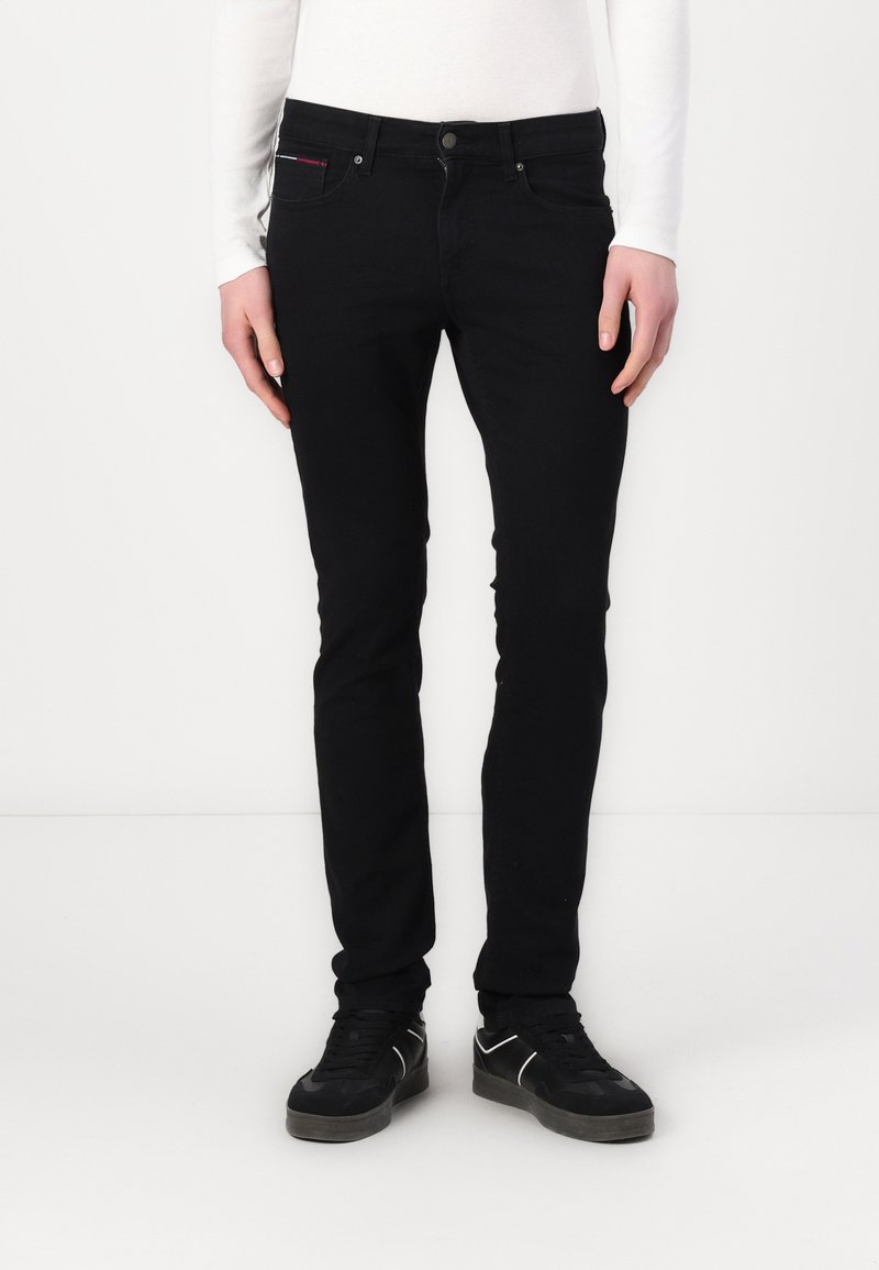 Persona vistiendo jeans negros ajustados y zapatillas negras, de pie contra un fondo blanco liso.