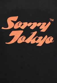 Πορτοκαλί στυλιζαρισμένο κείμενο "Sorry Tokyo" με σύμβολο TM σε μαύρο υφασμάτινο φόντο.