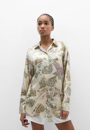 Jonge vrouw met ingevlochten haar, gekleed in een oversized overhemd met knopen, bedrukt met schetsen van beroemde Europese bezienswaardigheden op een lichte achtergrond.