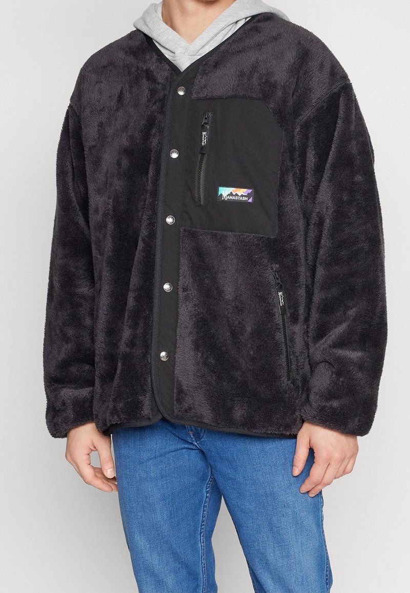 Persona che indossa una giacca nera pelosa con bottoni frontali, tasca laterale e tasca con zip sul petto, sopra un hoodie grigio chiaro e jeans blu.