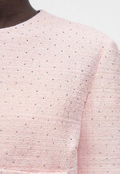 Gros plan d'une personne portant un haut texturé rose pâle orné de petites perles argentées réparties uniformément sur le tissu.