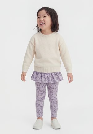 Enfant souriant debout, portant un pull crème, des leggings violets imprimés léopard avec une jupe à volants, et des chaussures blanches sans lacets.