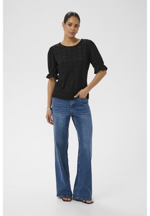 Žena oblečená v čiernej textúrovanej blúzke s nadýchanými rukávmi, modrých širokých džínsach a čiernych lodičkách s otvorenou špičkou, stojaca s jednou rukou v vrecku.