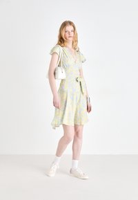 Abito giallo chiaro con motivo floreale, maniche corte a volant, vita con cintura e orlo svasato. Indossato con una borsa bianca e sneaker.