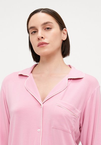 Light pink long-sleeve pajama top with a notched collar, front pocket, and contrasting white piping along the collar and button placket.