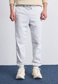 Lys grå joggebukser med elastisk linning og snor. Cuffede ankler, myk tekstur og en liten logo på låret. Kombinert med beige sneakers.