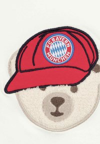 Red cap with FC Bayern München logo atop a plush bear face, featuring beige fur texture and embroidered facial details on a white background.