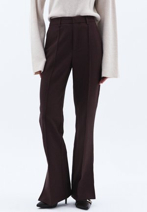 Pantaloni a zampa d'elefante marroni realizzati in tessuto strutturato. Presentano pieghe frontali e un alto punto vita. Abbinati a un maglione a coste color crema e tacchi neri.