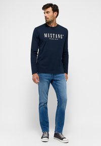 Hosszú ujjú sötétkék póló, fehér "MUSTANG" logóval, kék farmernadrággal és sötétkék sportcipővel párosítva. Sima pamut anyag, alakra szabott dizájnnal.