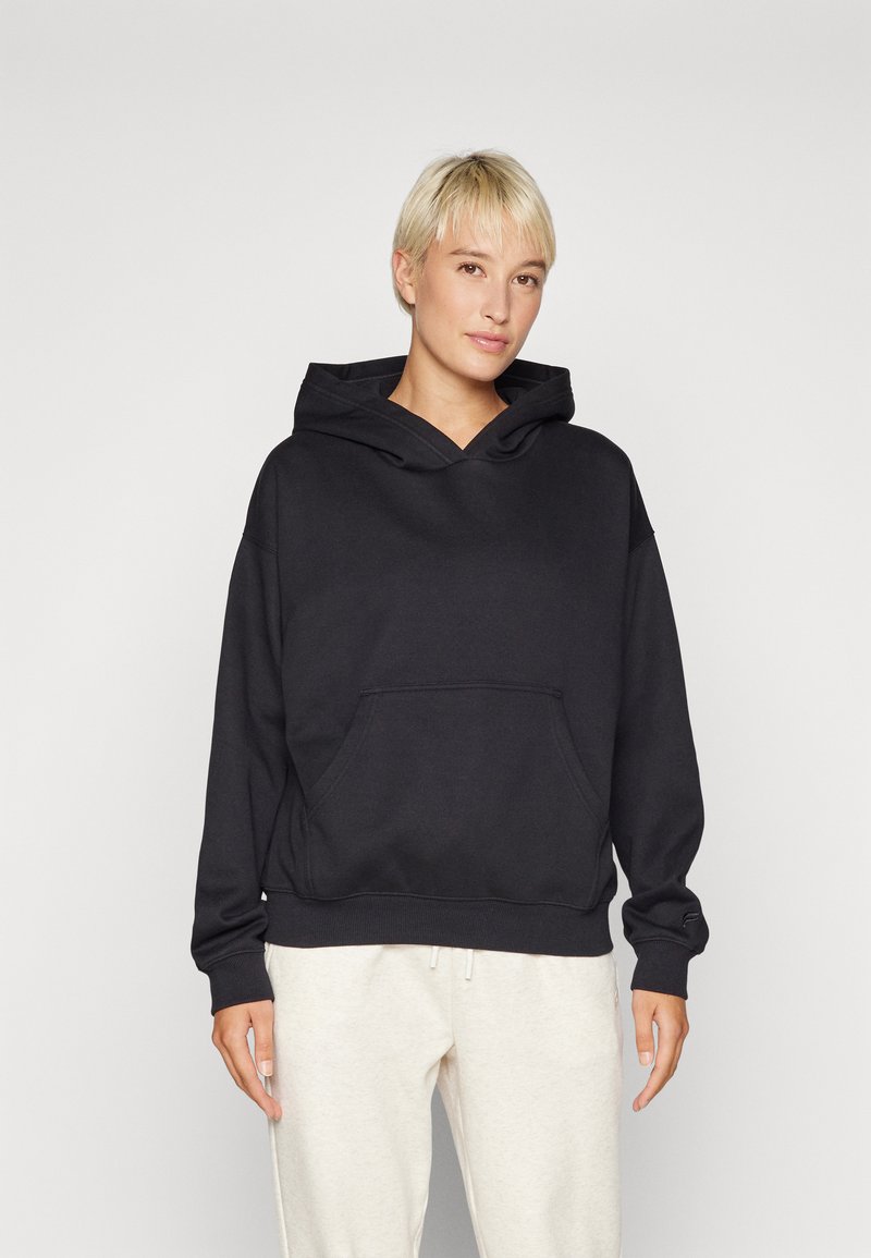 Felpa nera con cappuccio e tasca frontale, vestibilità comoda, maniche a polsino e una texture morbida, abbinata a pantaloni beige chiaro.