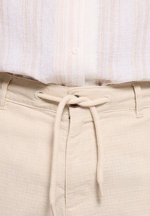 Primer plano de pantalones beige con un cordón atado en un nudo en la cintura, combinados con una camisa ligera abotonada.