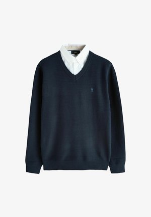 Un pull bleu marine en V superposé sur une chemise blanche à boutons, avec un petit logo brodé sur la poitrine gauche.