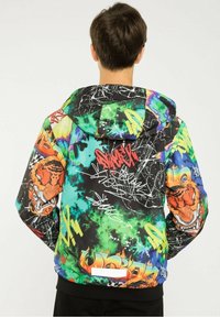 Personne portant une veste à capuche ornée de motifs de style graffiti colorés, comprenant des formes abstraites, des flammes et des visages de tigre sur les manches.