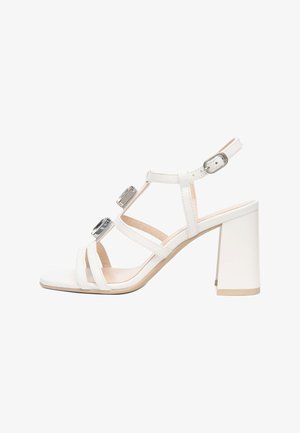 Sandale blanche à talon avec bride de cheville ajustable, deux grosses pierres rectangulaires sur les brides, et talon bloc sur fond blanc.