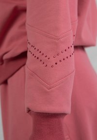 Der pinke Sweatshirtstoff verfügt über ein gemustertes Design aus kleinen roten Strasssteinen am Ärmel, mit einem gerippten Bund und Zickzacknähten.