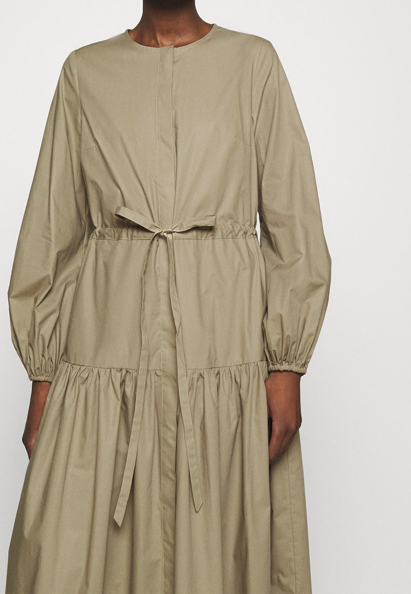 Femme portant une robe marron clair à manches longues avec taille froncée et ceinture à nouer, poignets élastiques et col rond sur un fond uni.