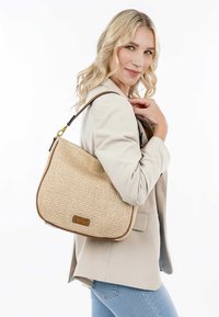 Sac à main en tissu naturel tressé avec une forme courbée, de couleur beige et avec des finitions marron. Équipé d'une bandoulière et d'un accent de logo à l'avant.